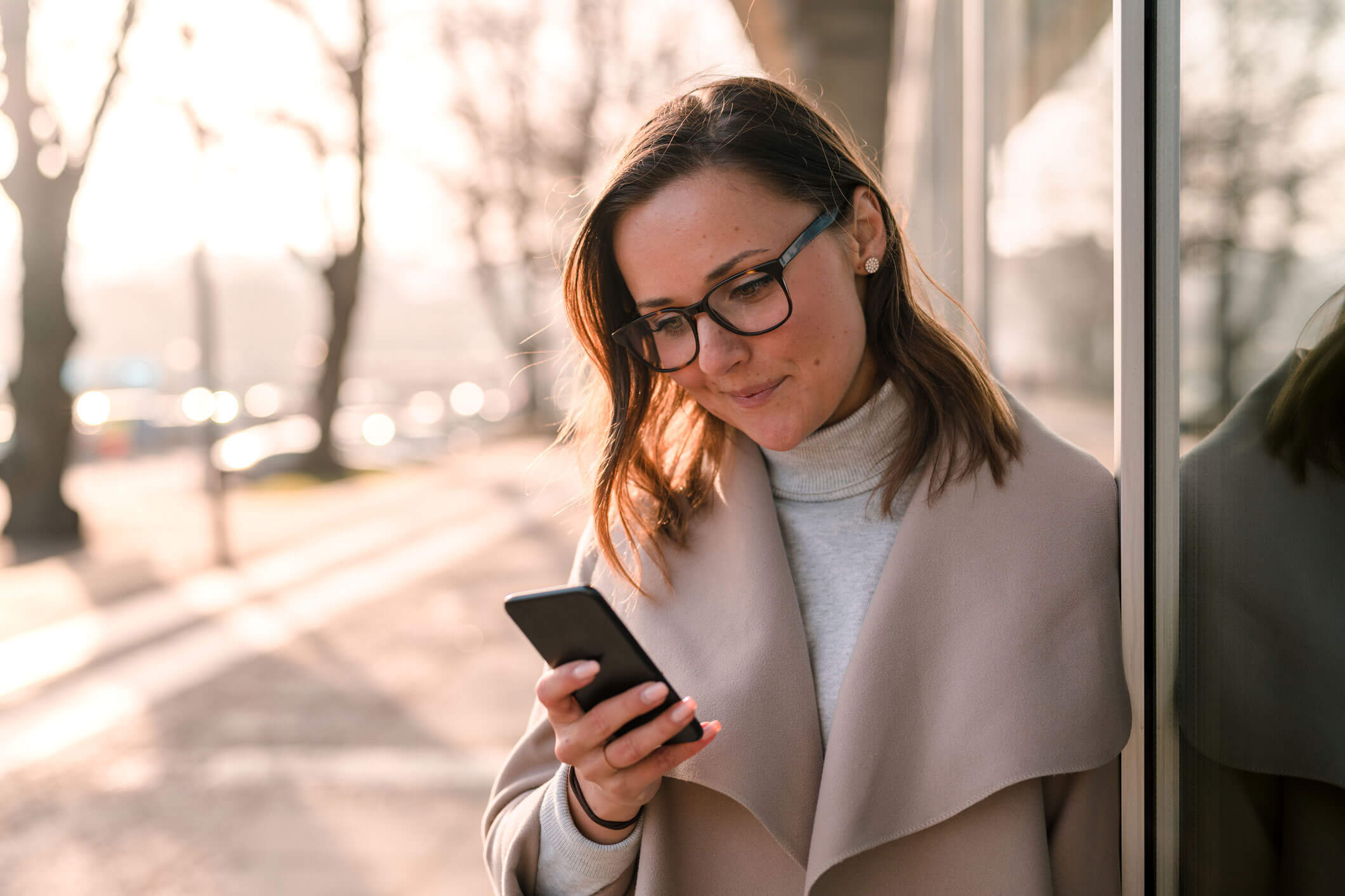 En kvinne står ute i vakkert høstvær og lener seg inntil en vegg mens hun ser på mobilen.