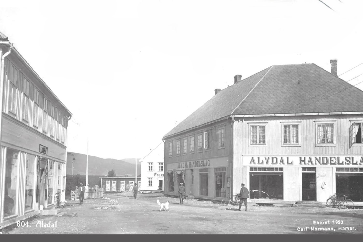 Bilde fra digitalt museum av Steia på Alvdal, Bildet viser en gate med folk som går, en hund og to store handelshus på hver side av gata, i bakgrunnen ser man folkets hus.