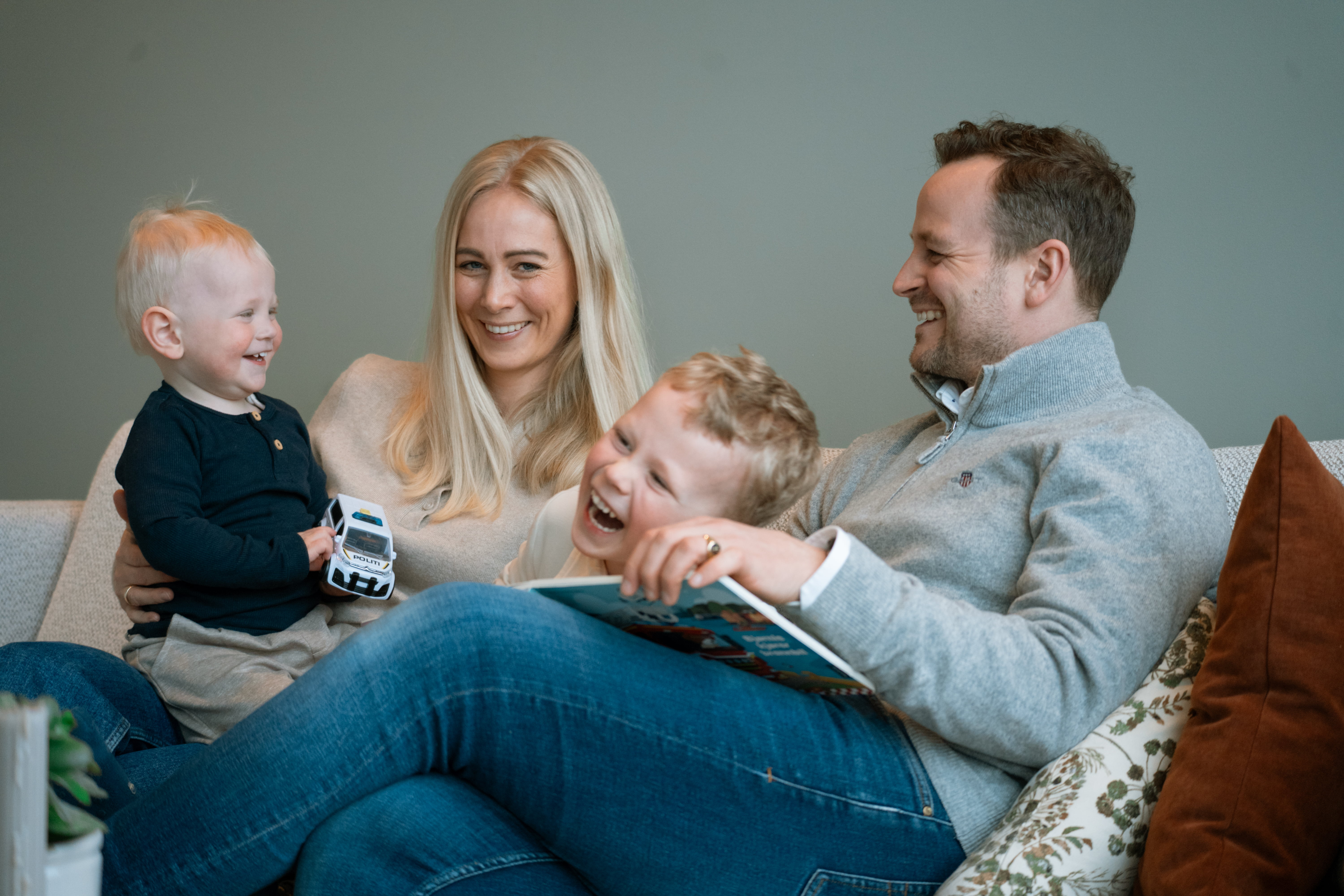 En familie sitter samlet i en sofa i en stue. En voksen holder et lite barn på fanget, mens et annet barn sitter mellom de voksne og ser i en bok. Barnet ytterst holder en liten lekebil. Sofaen har puter, og rommet har rolige farger og et hjemmekoselig preg.