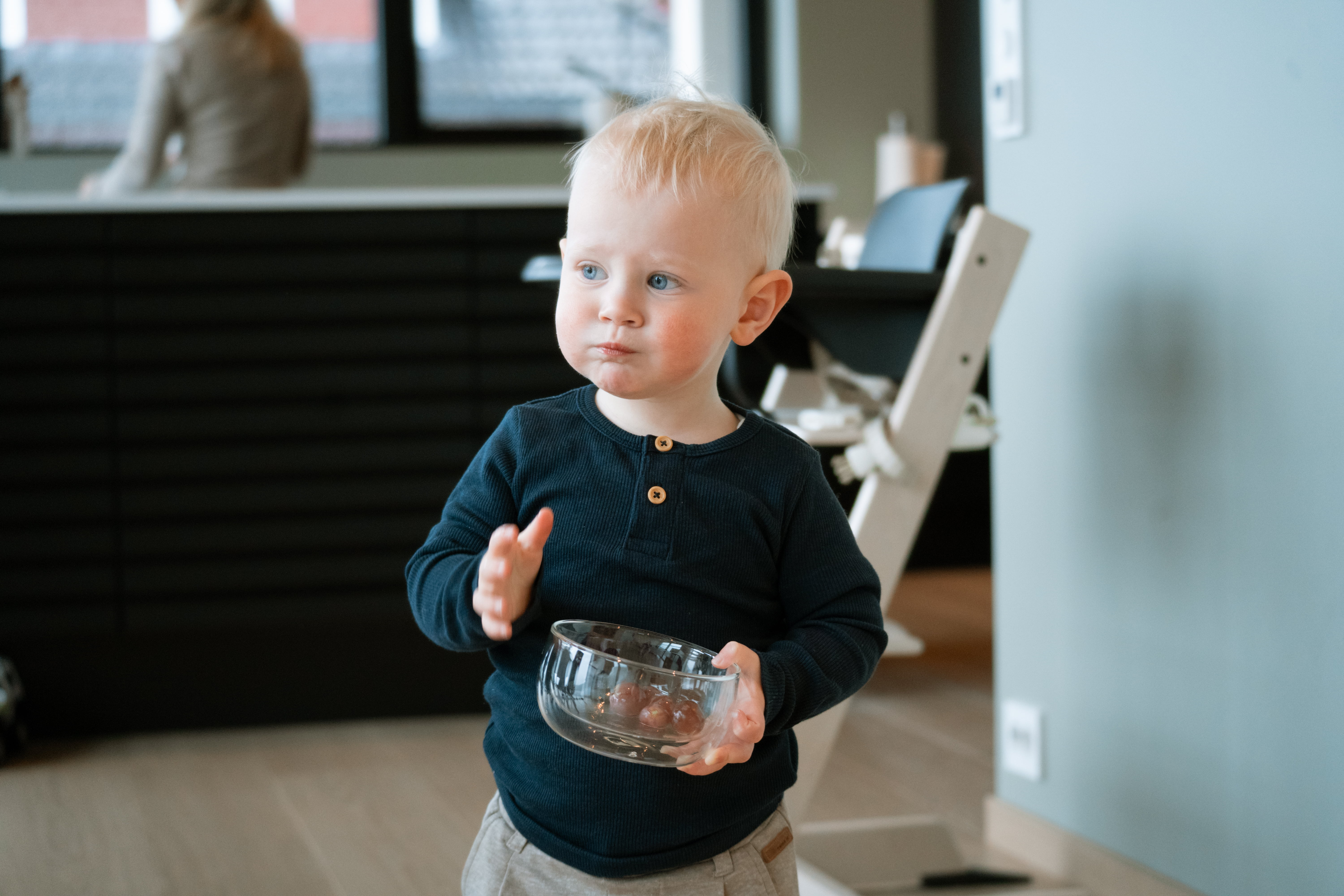 Et lite barn står innendørs og holder en gjennomsiktig bolle i hendene. Barnet har på seg en mørk langermet genser og lyse bukser. I bakgrunnen sees et kjøkken- og spiseområde med benk, stol og dempede farger.