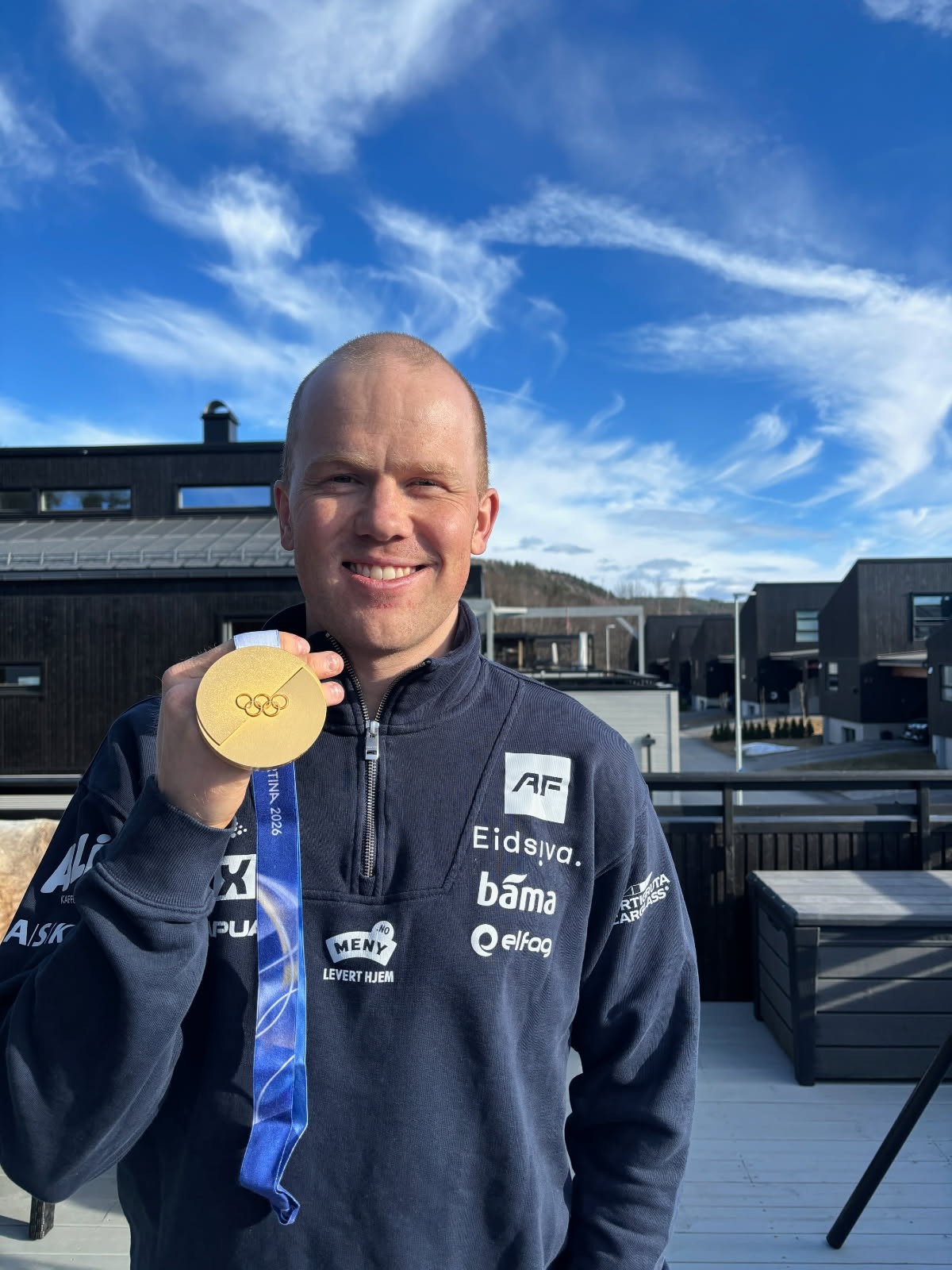 En person står utendørs på en terrasse og holder opp en gullfarget medalje i et blått bånd. Personen har på seg en mørk treningsjakke med flere sponsorlogoer. I bakgrunnen sees moderne trebygninger og en blå himmel med lette skyer.